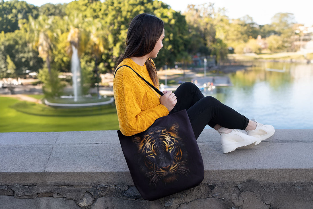 Woman sitting on a ledge in an outdoor park, wearing a bright yellow sweater and holding a black tote bag with a realistic tiger face design. A calm lake and a fountain are visible in the background, surrounded by lush green trees.
